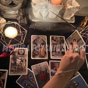 May include: Tarot cards with detailed illustrations, including one labelled "Death", are arranged on a black surface. A lit candle in a geometric holder and other decorative items are also present. The image has a mystical theme, with a hand holding a card.