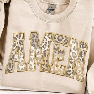 Può includere: Felpa beige con la scritta "AMEN" in stampa leopardata. Le lettere sono delineate da cuciture dorate. La felpa è realizzata in materiale morbido e ha uno scollo tondo.