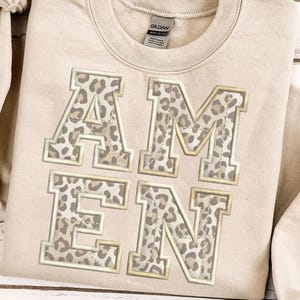 Può includere: Felpa beige con la scritta "AMEN" ricamata a grandi lettere con stampa leopardata e bordo dorato. La felpa ha un girocollo e un colore neutro, adatta a vari outfit.