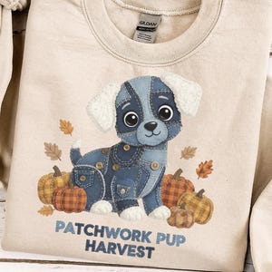 May include: Beige sweatshirt featuring a cartoon puppy made of denim patches, surrounded by pumpkins and fall leaves. The text "PATCHWORK PUP HARVEST" is printed below the puppy. The sweatshirt has a crew neck and long sleeves.