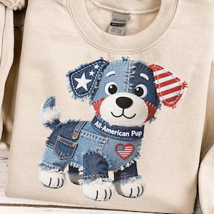 May include: A beige sweatshirt featuring a patchwork puppy design. The puppy has denim overalls, a blue and red head, and a collar that reads "All-American Pup." The design incorporates stars and stripes.