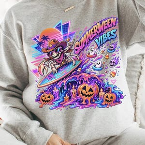 May include: A light gray sweatshirt with a colorful graphic. The graphic features a skeleton cowboy surfing a wave with pumpkins, ghosts, and the text "SUMMERWEEN VIBES". The design includes a sunset and geometric shapes.