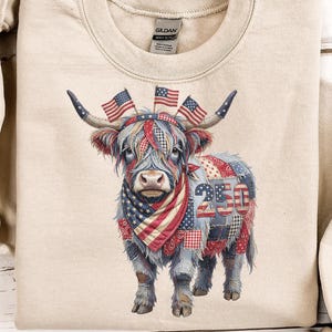 Può includere: Felpa beige con un design di mucca delle Highlands. La mucca è adornata con motivi della bandiera americana, tra cui una bandana, patchwork e bandiere sulle corna. Il numero "250" è visualizzato sul lato della mucca.