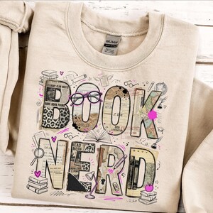 May include: Beige sweatshirt with a graphic that says "BOOK NERD". The letters are designed with book pages, leopard print, and pink accents. The design includes glasses, books, and a lamp.