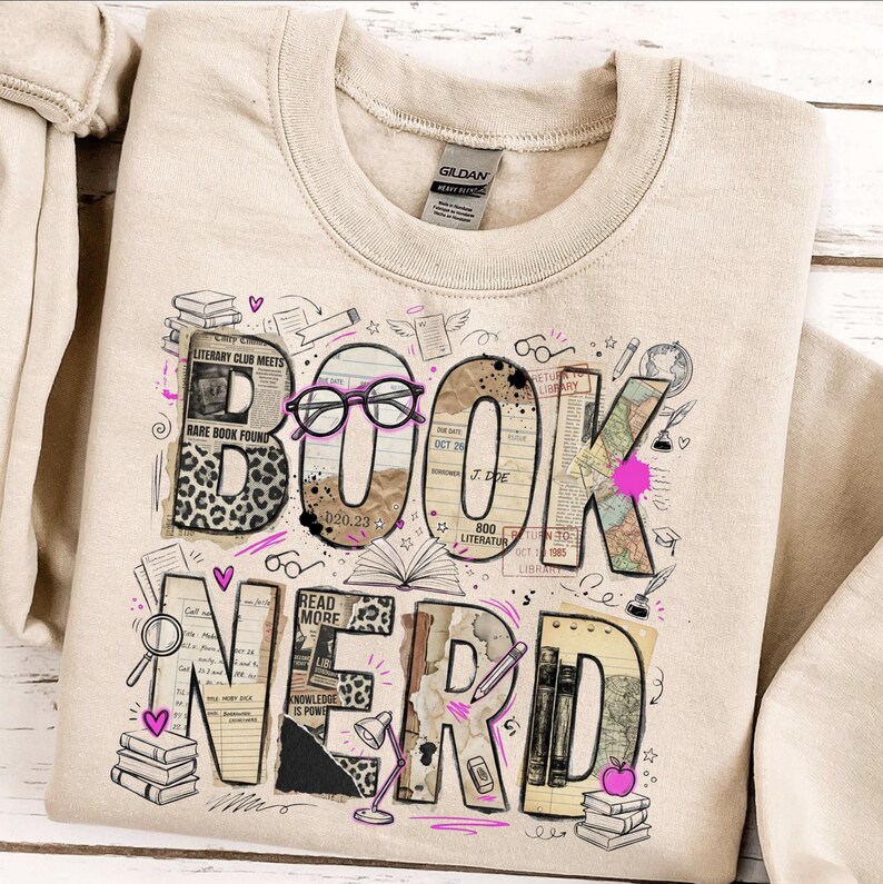 May include: Beige sweatshirt with a graphic that says "BOOK NERD". The letters are designed with book pages, leopard print, and pink accents. The design includes glasses, books, and a lamp.