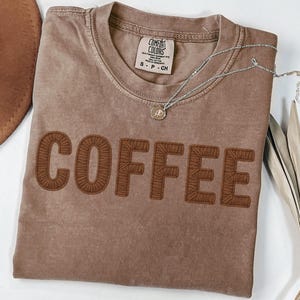 Puede incluir: Una camiseta marr&oacute;n Comfort Colors con la palabra "COFFEE" bordada en un marr&oacute;n m&aacute;s oscuro. Un collar plateado con un peque&ntilde;o colgante descansa sobre la camisa. La camisa est&aacute; doblada sobre una superficie blanca.