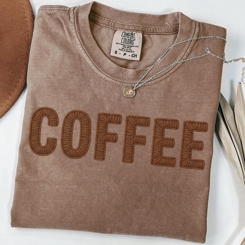 Puede incluir: Una camiseta marr&oacute;n Comfort Colors con la palabra "COFFEE" bordada en un marr&oacute;n m&aacute;s oscuro. Un collar plateado con un peque&ntilde;o colgante descansa sobre la camisa. La camisa est&aacute; doblada sobre una superficie blanca.