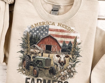 Könnte beinhalten: Beiges Sweatshirt mit einer Grafik, die eine rote Scheune, die amerikanische Flagge, einen Traktor, eine Kuh und den Text "AMERICA NEEDS FARMERS" zeigt. Das Design umfasst Baumwolle und Maisstängel und schafft ein ländliches Thema.