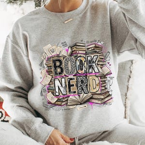 Puede incluir: Una sudadera gris claro con las palabras "BOOK NERD" en un dise&ntilde;o apilado. Las letras tienen un estampado de leopardo y un contorno negro. El dise&ntilde;o incluye pilas de libros, libros abiertos y las palabras "Read More" y "Librarian Vibes".