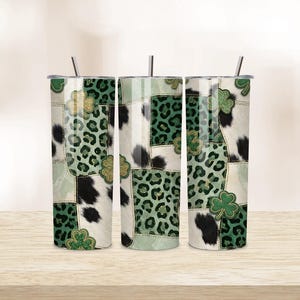 May include: Three stainless steel tumblers with lids and straws. The tumblers feature a patchwork design with cowhide, leopard print, and shamrock accents in green, black, and gold. The tumblers are cylindrical and sit on a wooden surface.