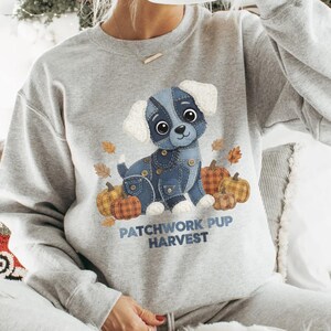 Puede incluir: Sudadera gris claro con un cachorro de dibujos animados hecho de parches de mezclilla, rodeado de calabazas y hojas de oto&ntilde;o. El texto "PATCHWORK PUP HARVEST" est&aacute; impreso debajo.