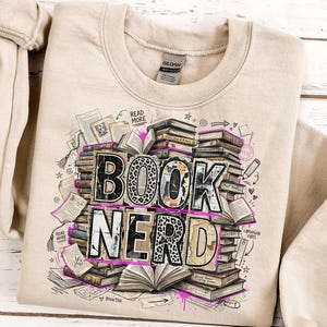 Puede incluir: Sudadera beige con las palabras "BOOK NERD" en un dise&ntilde;o apilado. Las letras tienen un estampado de leopardo y est&aacute;n rodeadas de ilustraciones de libros, p&aacute;ginas abiertas y el texto "READ MORE".