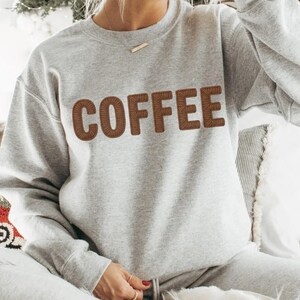 Puede incluir: Una sudadera gris claro con la palabra "COFFEE" bordada en letras marrones. La sudadera forma parte de un conjunto a juego. El dise&ntilde;o es sencillo e informal, adecuado para el uso diario. El material parece suave.