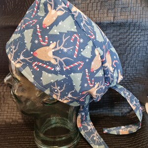 Könnte beinhalten: Eine marineblaue OP-Haube mit einem festlichen Weihnachtsmuster. Das Design umfasst Rentiere, Weihnachtsbäume, Zuckerstangen und Schneeflocken. Die Haube hat Bänder für eine verstellbare Passform.