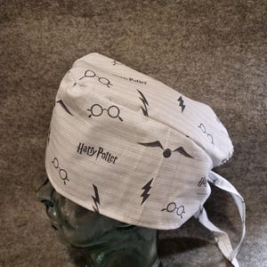Könnte beinhalten: Eine weiß-grau gemusterte Mütze mit den Worten "Harry Potter", Brillen, Blitzsymbolen und einem geflügelten goldenen Schnatz. Die Mütze hat einen Bindeband am Rücken und ist aus weichem Stoff gefertigt.