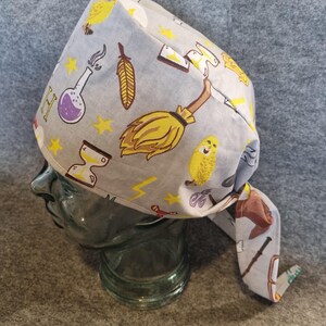 Könnte beinhalten: Eine graue OP-Haube mit einem sich wiederholenden Muster aus gelben Sternen, einem Besen, einer Eule, einer Feder und Zaubertränken. Die Haube ist am Rücken gebunden und wird auf einem Glaskopf präsentiert.