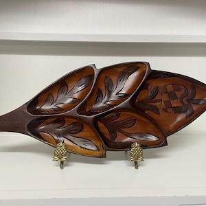 May include: A carved wooden leaf-shaped serving tray with multiple compartments. The tray is made of dark brown wood with lighter brown accents and features carved leaf and pineapple designs. It is supported by two brass pineapple-shaped stands.
