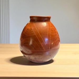 May include: A handcrafted, reddish-brown ceramic vase with a rounded body and a narrow neck. The vase features etched geometric patterns, including curved lines and triangles. The vase is displayed on a light-colored wooden surface.