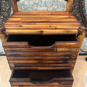 May include: A bamboo storage chest with two pull-out drawers and a curved handle. The chest is constructed from horizontal bamboo slats, showcasing natural wood tones. The drawers have curved cutouts for easy access, and the overall design is rustic and functional.