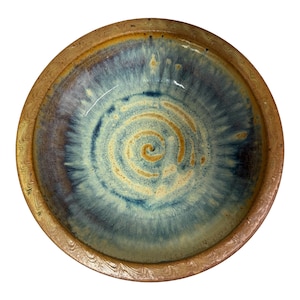 May include: A round ceramic bowl featuring a wide, textured tan rim with a wavy pattern. The interior displays a swirling glaze effect in shades of deep blue, light blue, and cream, radiating from a central point to the bowl's edge.