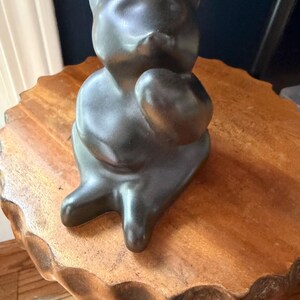 May include: A smooth, matte black ceramic figurine shaped like a seated bear. The bear is depicted in a stylized, rounded form with its head tilted slightly upward and one paw raised toward its chest, featuring a solid dark finish throughout.