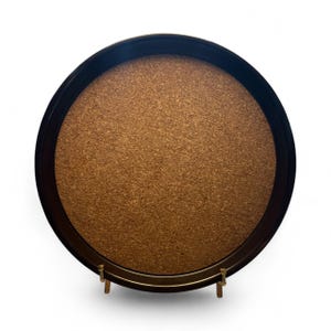 May include: A round serving tray with a dark brown rim and a cork center. The tray is supported by a gold-colored metal stand. The tray is designed for serving food or drinks.