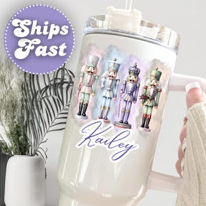 May include: White tumbler featuring a design of four toy nutcracker soldiers in various colors. The tumbler has a handle and a straw. The name "Kailey" is written in cursive. A purple circle with the words "Ships Fast" is also visible.