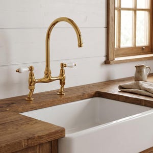May include: A polished gold kitchen tap with a high-arc spout and two white ceramic lever handles. The tap is mounted on a wooden worktop next to a white farmhouse sink, featuring a classic bridge design with a metallic gold finish.