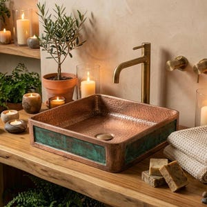 Könnte beinhalten: Ein kupfernes Waschbecken mit quadratischem Becken und grünem Patinarand steht auf einer Holzplatte. Eine goldene Armatur und Wandbefestigungen sind installiert. Die Badezimmereinrichtung umfasst Kerzen, Pflanzen und Handtücher.