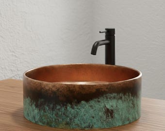May include: A round copper sink with a dark green and brown patina. The interior of the sink is a bright copper color. A black faucet is mounted on the wall behind the sink. The sink is sitting on a wooden surface.