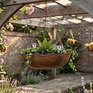 May include: A hanging copper-colored planter filled with greenery and purple and white pansies. The planter is suspended from a wooden pergola with climbing roses and other hanging baskets in the background. The scene is outdoors.