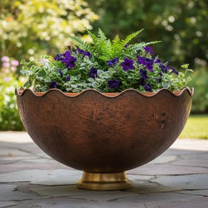Puede incluir: Una gran jardinera de cobre martillado con un borde festoneado, llena de vibrantes petunias moradas y follaje verde. La jardinera se asienta sobre una base corta de color dorado, perfecta para la decoración exterior. El fondo está borroso, sugiriendo un jardín.