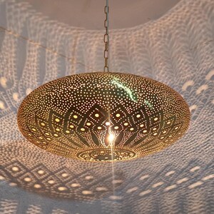 May include: A gold-colored, pendant light fixture with a perforated design, hanging from a chain. The light casts intricate patterns on the surrounding surface. The fixture has a round, flattened shape and a warm glow.