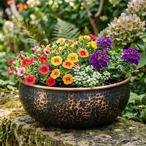 Könnte beinhalten: Eine bronzefarbene, gehämmert-metallene Pflanzschale, die mit bunten Blumen überquillt. Die Blumen umfassen rote, rosa, gelbe, orangefarbene, weiße und violette Blüten. Die Schale steht auf einer moosigen Steinmauer, umgeben von Grün.