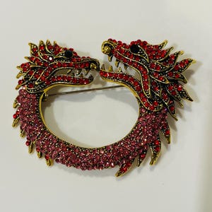 May include: A gold-toned dragon brooch with red and pink rhinestones. The brooch features two dragon heads facing each other, forming a circular shape. The dragons have intricate details and black eyes.
