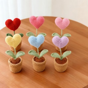 Könnte beinhalten: Sechs gehäkelte herzförmige Blumen in kleinen Töpfen. Die Herzen sind rot, rosa, gelb, blau und lila. Die Töpfe sind braun und beige, mit grünen Blättern. Die Blumen sind auf einer Holzoberfläche angeordnet.