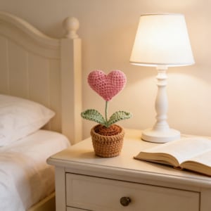 Puede incluir: Una flor de ganchillo con forma de corazón rosa, hojas verdes y un tallo marrón, plantada en una maceta marrón pequeña. La maceta está sobre una mesita de noche blanca, junto a una lámpara y un libro abierto.