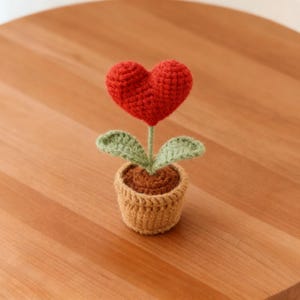 Puede incluir: Una flor de ganchillo con un corazón rojo, hojas verdes y una maceta beige llena de 'tierra' marrón. La flor está sobre una superficie de madera. Un objeto artesanal encantador.