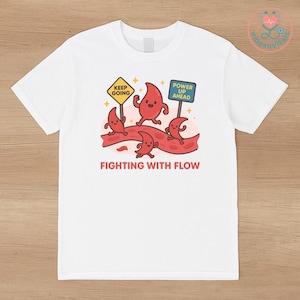 May include: White t-shirt featuring a cartoon design of red blood cells running on a red vein. The design includes signs that say "Keep Going" and "Power Up Ahead" with the text "Fighting With Flow" below.