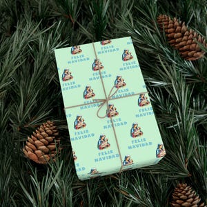 May include: A wrapped gift with light green paper featuring a repeating pattern of a religious image and the words "Feliz Navidad" in blue. The gift is tied with twine and set against a backdrop of pine needles and pine cones.