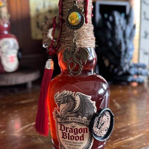 Peut inclure: Une bouteille décorative en verre remplie de liquide rouge, étiquetée "Dragon Blood" d'Arcane Apothecary. La bouteille est ornée d'un charme de dragon en bronze, d'un gland rouge et d'un sceau de cire. Le dessus est enveloppé de ficelle et scellé avec de la cire rouge.