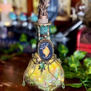 May include: A decorative glass bottle featuring a bronze fairy figurine. The bottle has a teal and brown design with a label reading "Will O' The Wisps". It's embellished with chains and a green gem, creating a whimsical aesthetic.