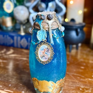 May include: A glass bottle filled with shimmering blue liquid, featuring a gold-painted base and a cork stopper. The neck is wrapped in twine with gold wax drips, adorned with a gold-framed oval pendant depicting a figure and a small leaf charm.