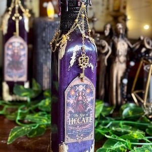 May include: A decorative purple bottle with gold accents and a black neck, adorned with a gold key charm and chain. The label reads "The Keys of Hecate" with an illustration of a goddess. The bottle has a textured, aged appearance.