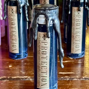 May include: Dark blue glass bottle with a silver wax seal and a label that reads "PROTECTION". The bottle contains a blue liquid with glitter. The label also includes the text "Hold in hand, set forth your intention (be specific), shake to activate."