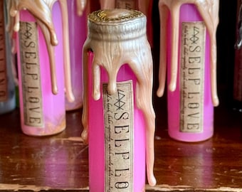 May include: Pink glass bottles with gold-colored wax drippings. Each bottle has a label that reads "SELF LOVE" with additional text. The bottles are filled with a pink liquid and a golden substance at the bottom.