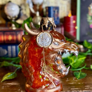 May include: A decorative bottle with a dragon head design. The bottle is primarily orange and gold, with intricate details and a coin-shaped charm hanging from the neck. The dragon's head features horns and sharp teeth, creating a fantasy theme.