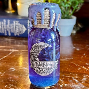 May include: A small glass bottle filled with a shimmering purple liquid, labelled "Wolfsbane." The bottle is sealed with a twine-wrapped cork and a dripped, iridescent purple wax. The bottle has a blue and purple gradient.