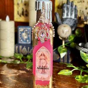 May include: A decorative glass bottle with a pink and gold label featuring an illustration of a woman. The bottle is adorned with a jeweled pendant and a light blue wax top. The label reads "The Passion of Aphrodite".