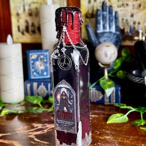 May include: A dark purple bottle with a red wax-sealed top and silver chain detail. The label features an illustration of a woman and the text "The Shadows of Lilith". The bottom of the bottle has a silver metallic finish.
